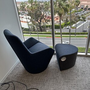Grey swivel chair with ottoman in modern office corner, overlooking cityscape through large windows.