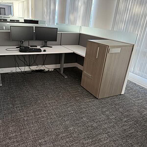 Office cubicle with dual monitors and wooden storage cabinet.