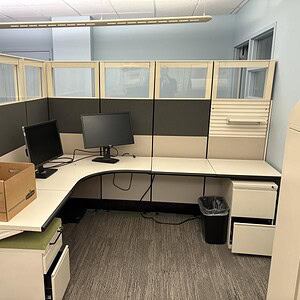 Office cubicles with beige and black panels, dual monitors, and storage drawers.