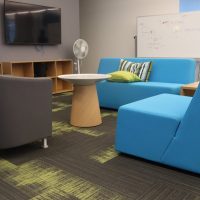 Bright lounge with blue and gray chairs, whiteboard, and TV screens for collaborative work.