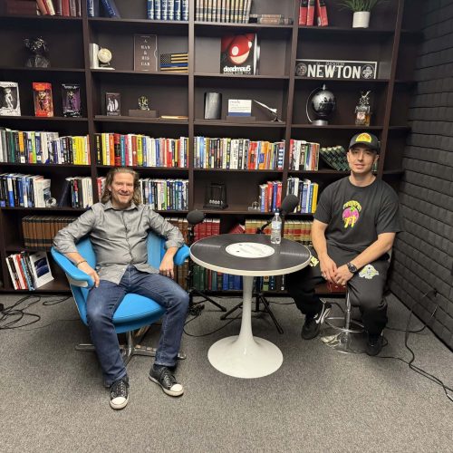 Two men seated at a table in a modern office with bookshelves, discussing business.