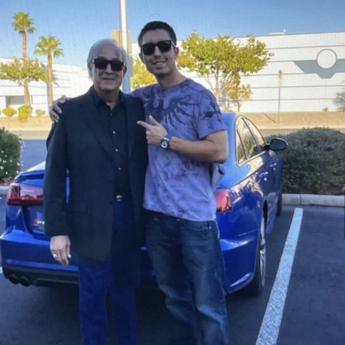 Two men standing next to a blue car in a parking lot, smiling.