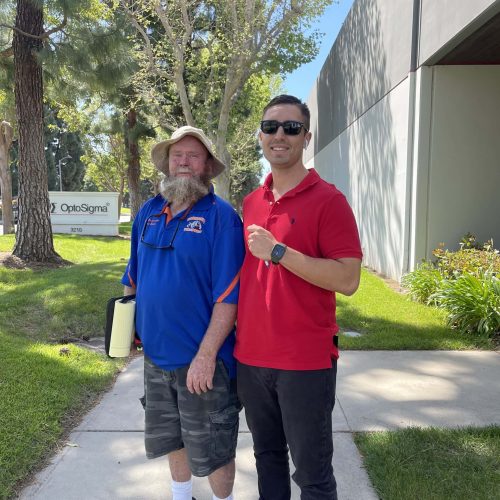 Two men standing outdoors, smiling, in front of a modern building and trees.
